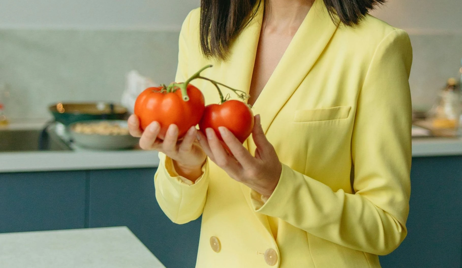 Frau in gelbem Blazer hält zwei frische Tomaten in der Küche.