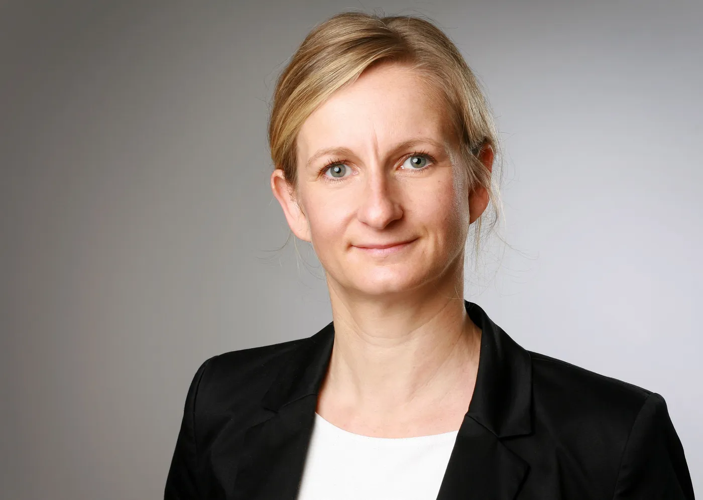 Porträt einer Frau mit blonden Haaren, die in einem schwarzen Blazer lächelt.