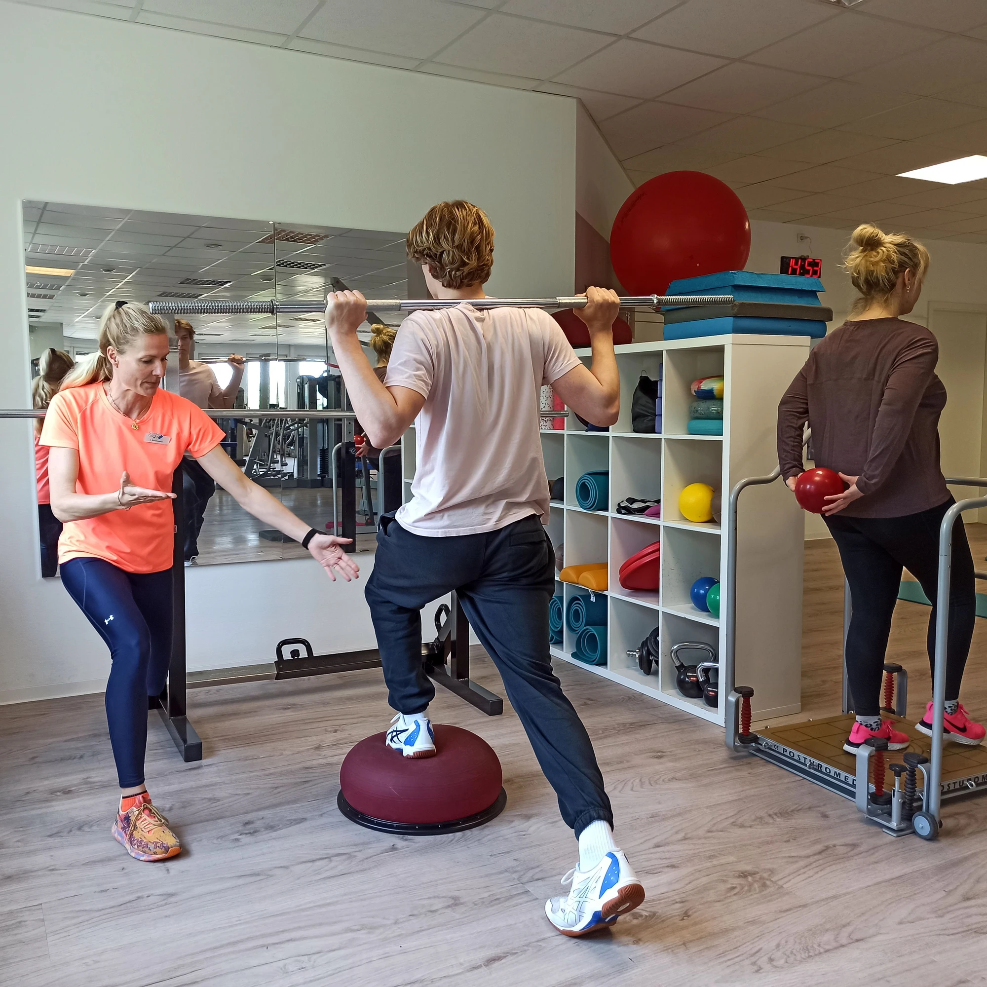 Eine Trainerin gibt Anweisungen, während ein Mann auf einem Balance-Pad trainiert.