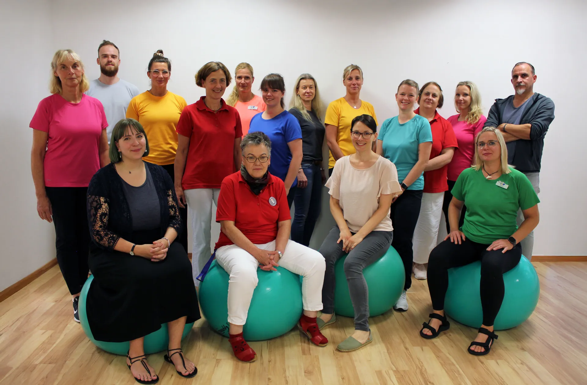 Gruppe von 18 Personen in bunten T-Shirts, sitzend und stehend auf Gymnastikbällen.