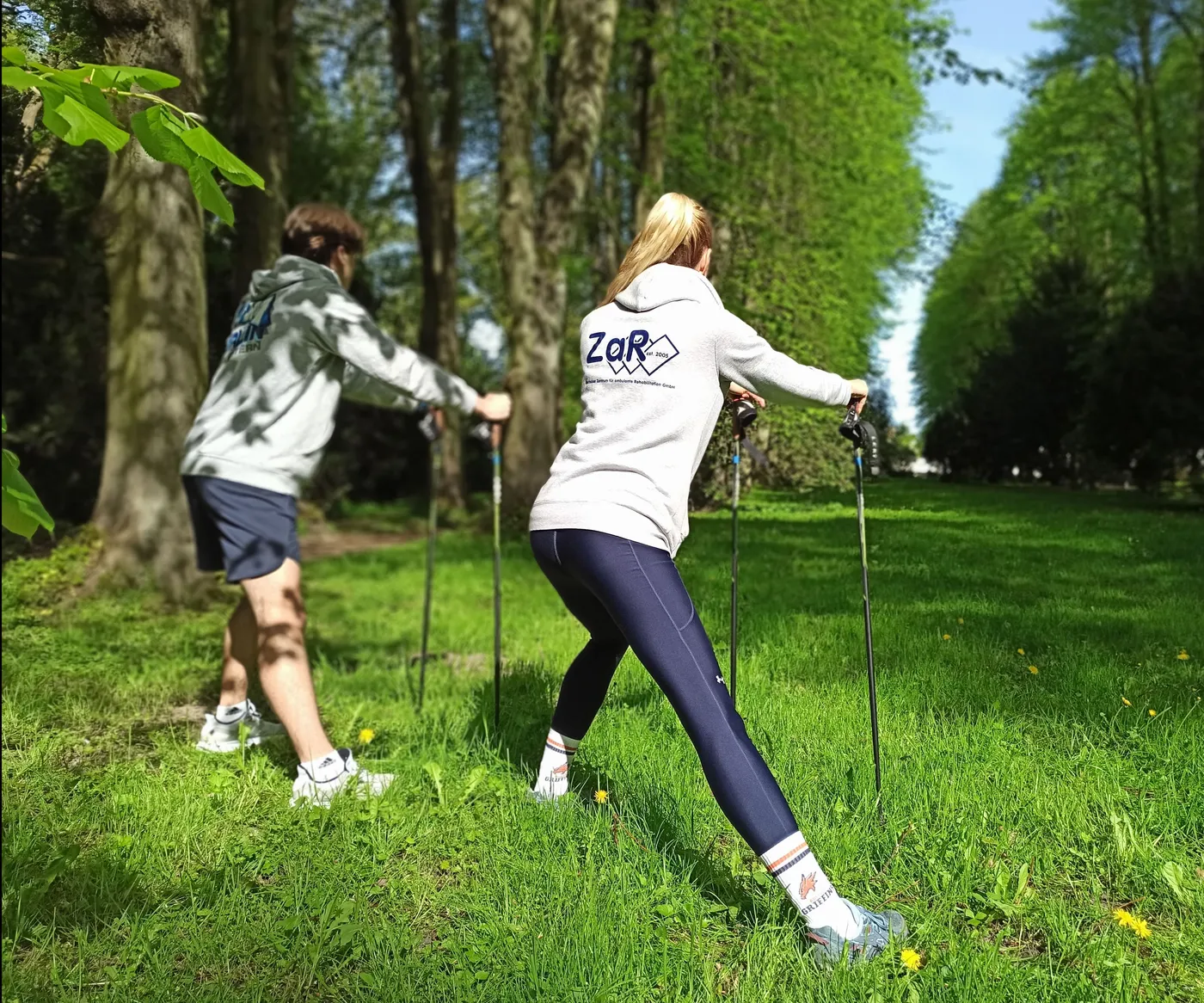 Zwei Personen beim Nordic Walking in einem grünen Park mit Bäumen.