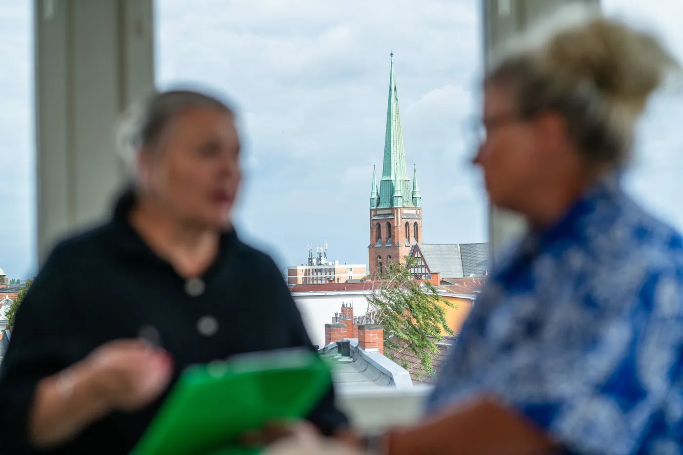 Zwei Frauen diskutieren, im Hintergrund ist ein Kirchturm sichtbar.