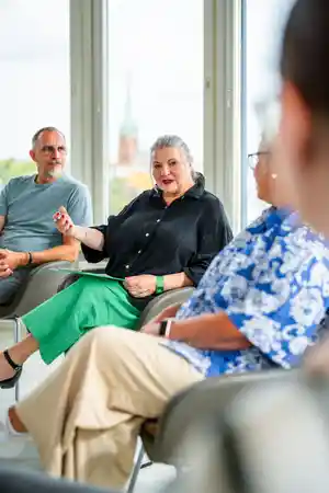 Eine Frau in schwarzer Bluse und grüner Hose spricht lebhaft mit Zuhörern in einem hellen Raum.