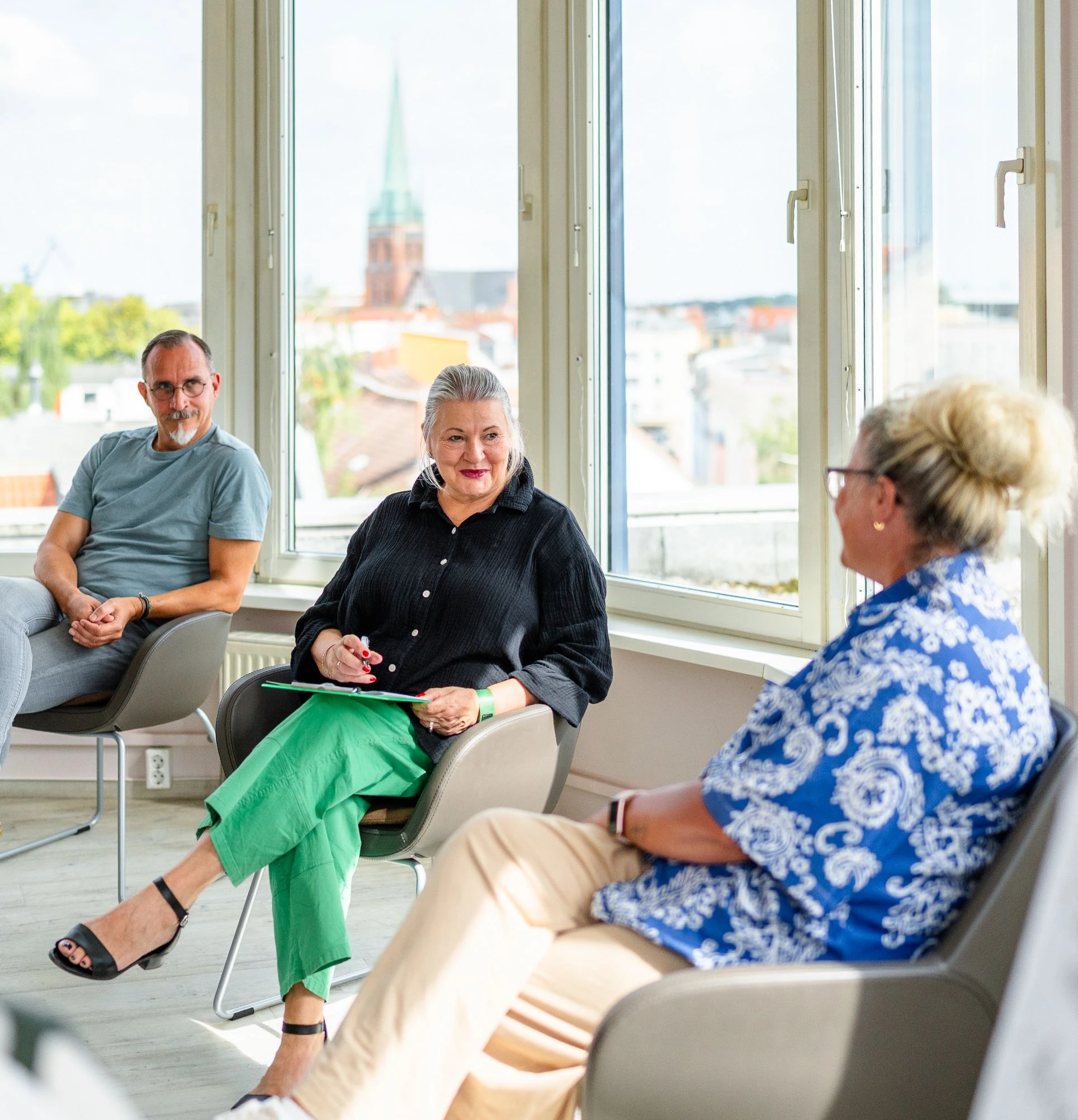 Drei Personen in einem hellen Raum diskutieren, mit Blick auf die Stadt durch große Fenster.