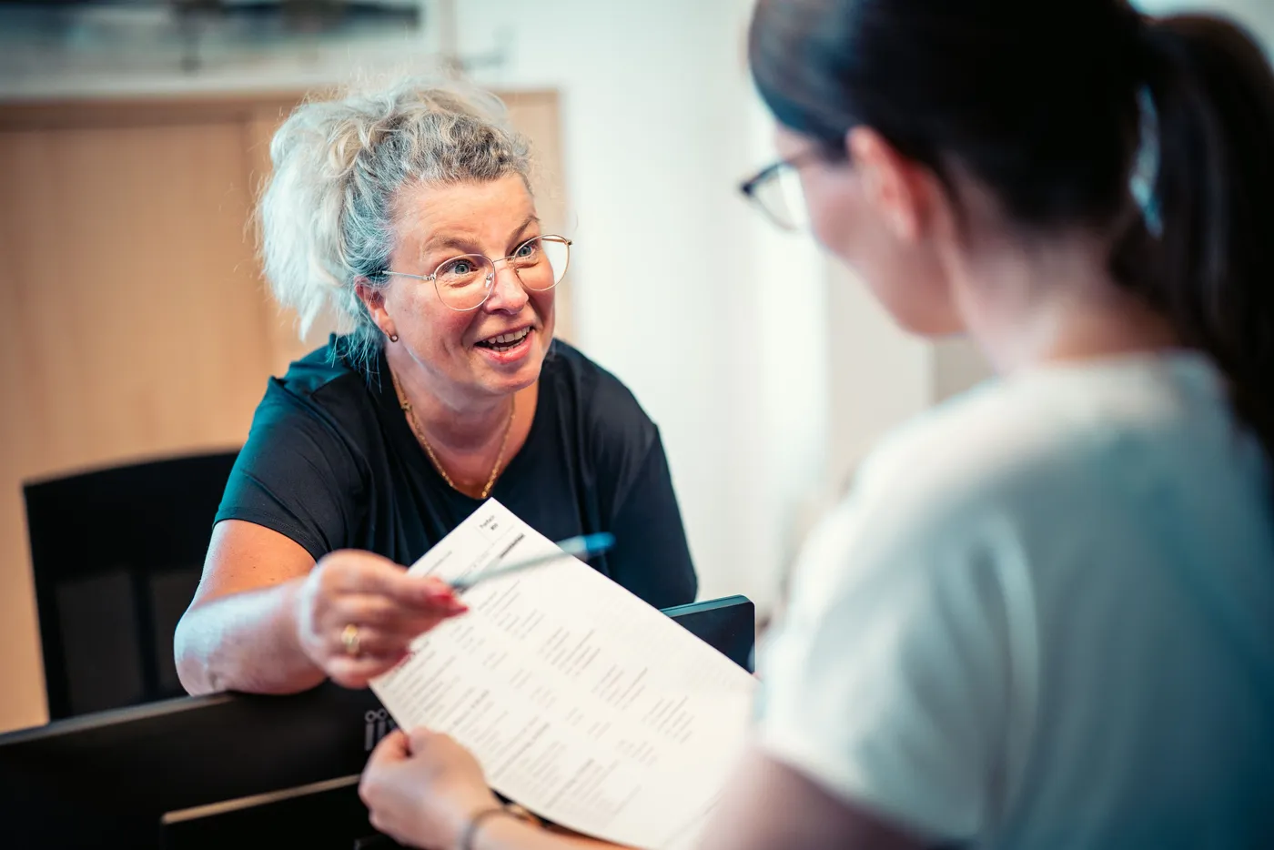 Eine Frau am Empfang lächelt und erklärt etwas zu einem Dokument an eine andere Person.