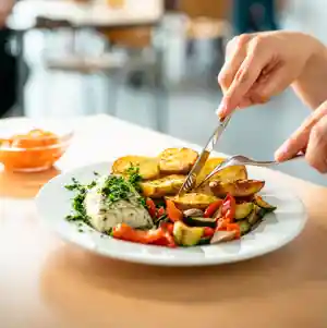 Eine Hand schneidet gebratene Kartoffeln und Gemüse auf einem Teller mit Fisch.
