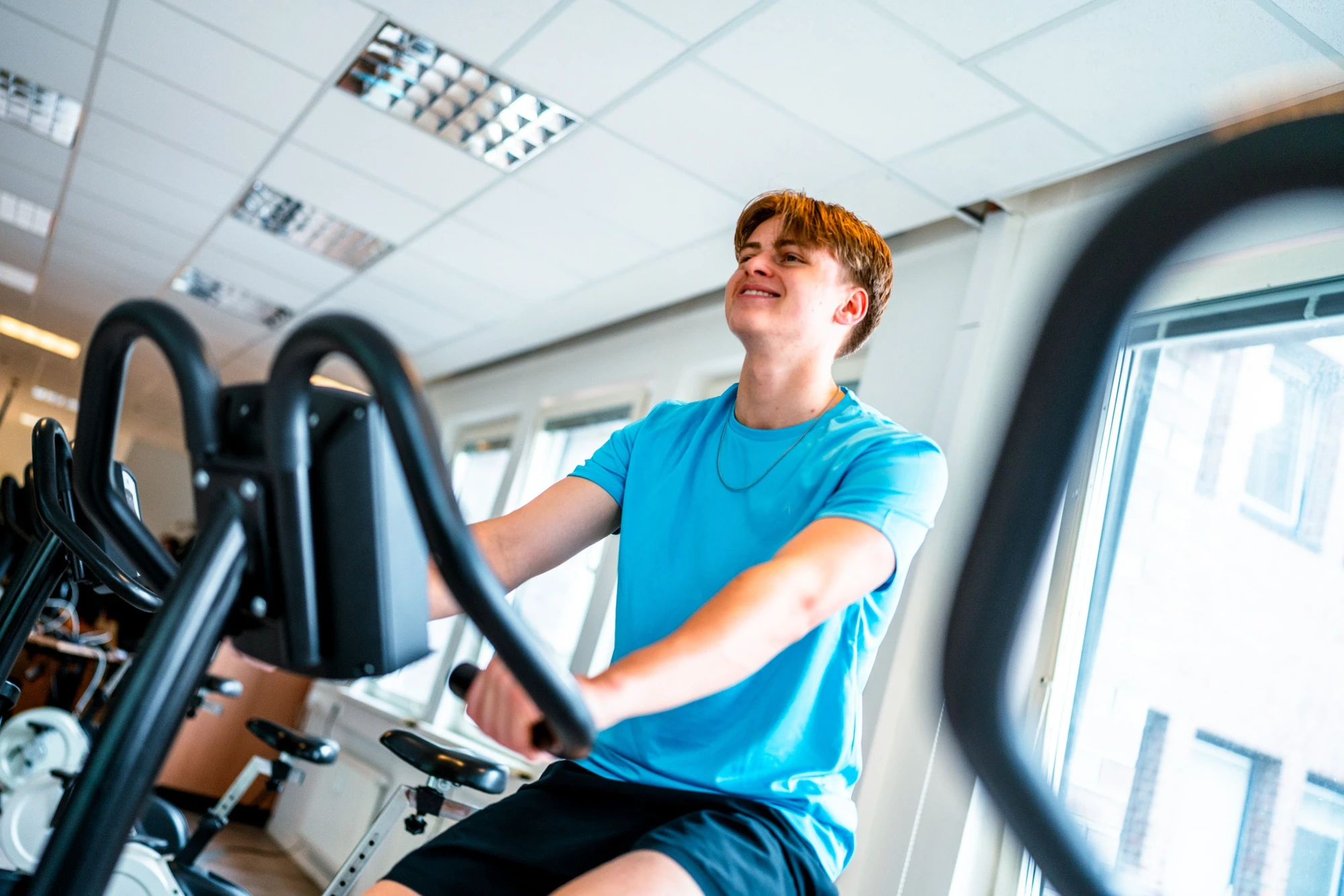 Junger Mann trainiert lächelnd auf einem Fitnessgerät in einem hellen Raum.