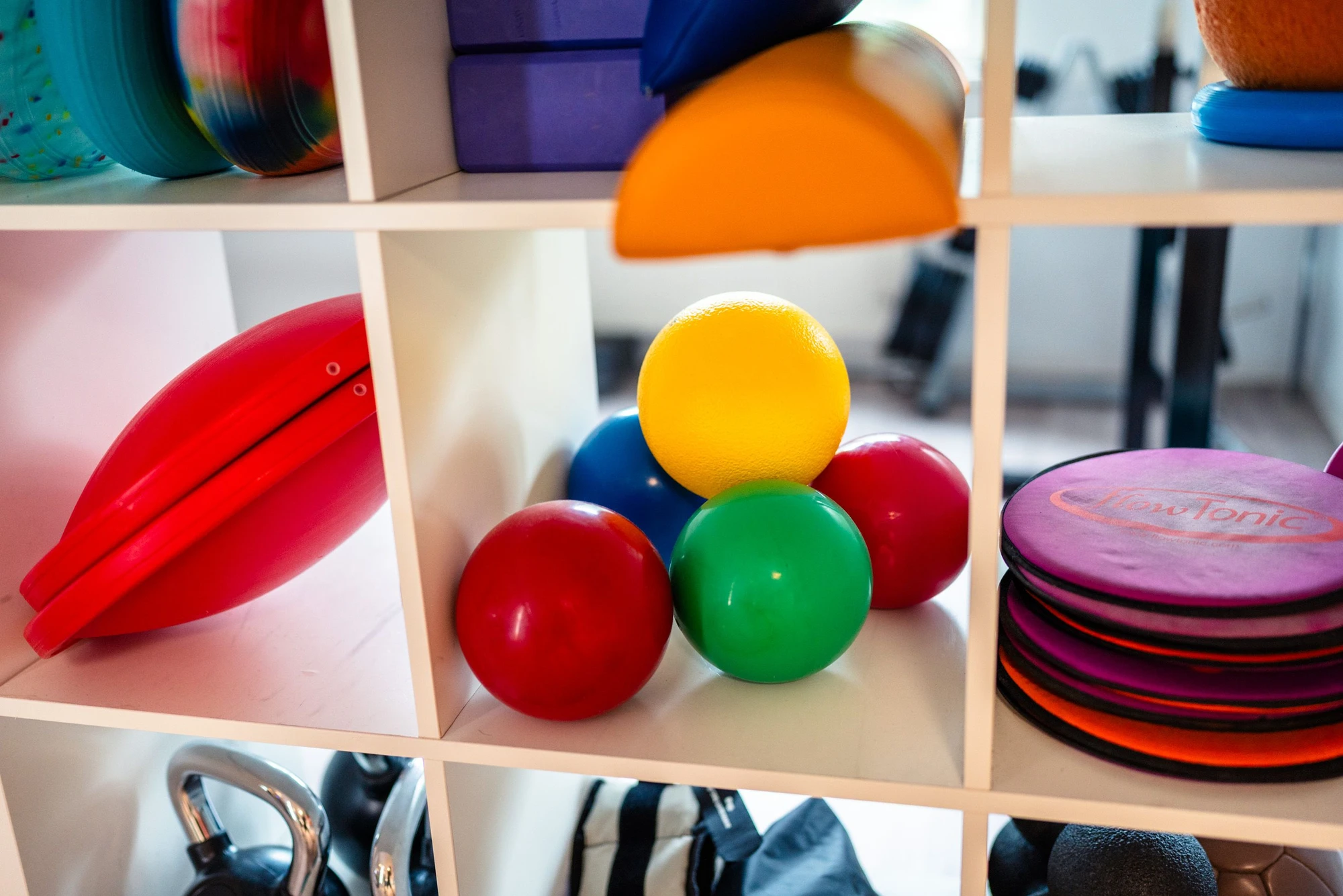 Bunte Bälle und Fitnessgeräte auf einem Regal in einem Trainingsraum.
