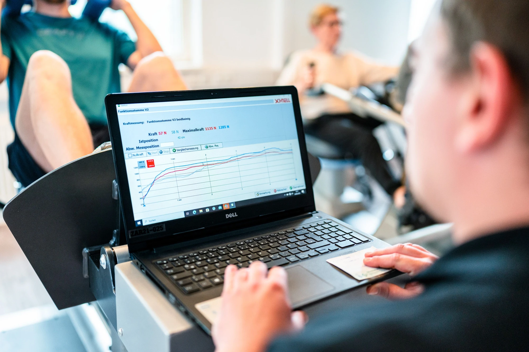Junge analysiert Trainingsdaten auf Laptop während einer Fitnessübung im Hintergrund.