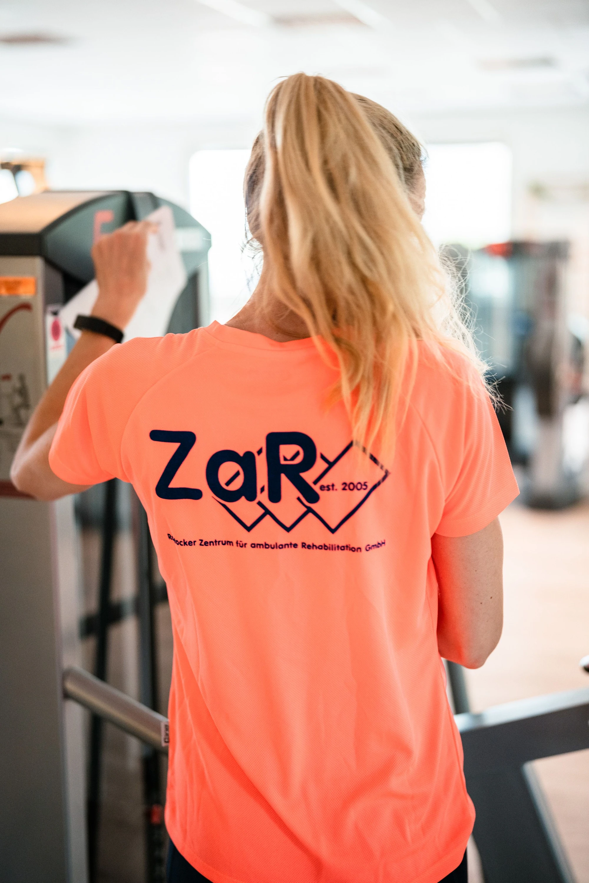 Eine Person in einem leuchtend orangefarbenen T-Shirt reinigt ein Fitnessgerät.