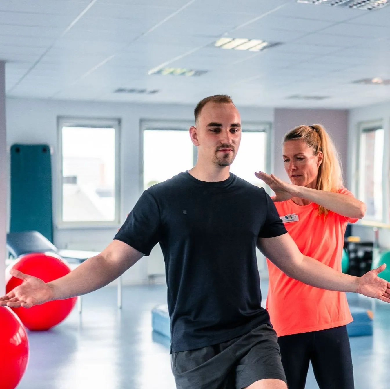 Ein Mann balanciert mit Unterstützung einer Trainerin in einem Fitnessraum.
