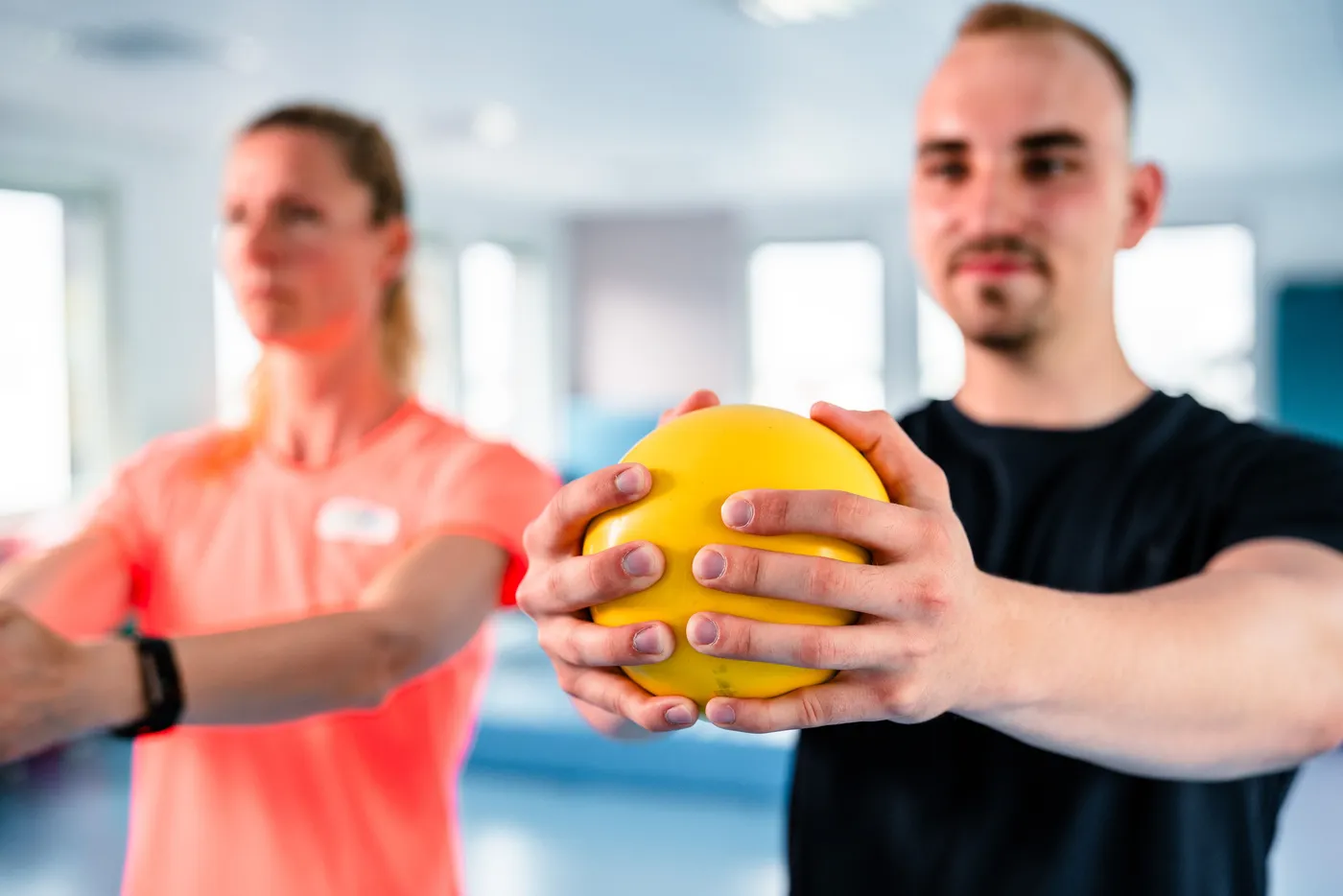 Zwei Personen halten einen gelben Ball, während sie in einem Fitnessraum trainieren.