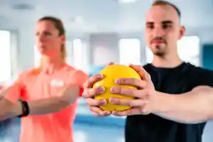 Zwei Personen halten einen gelben Ball, während sie in einem Fitnessraum trainieren.
