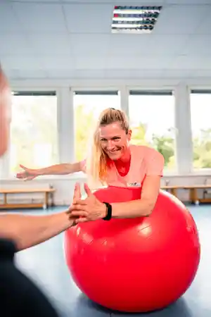 Frau auf einem Gymnastikball streckt die Arme aus und lächelt.