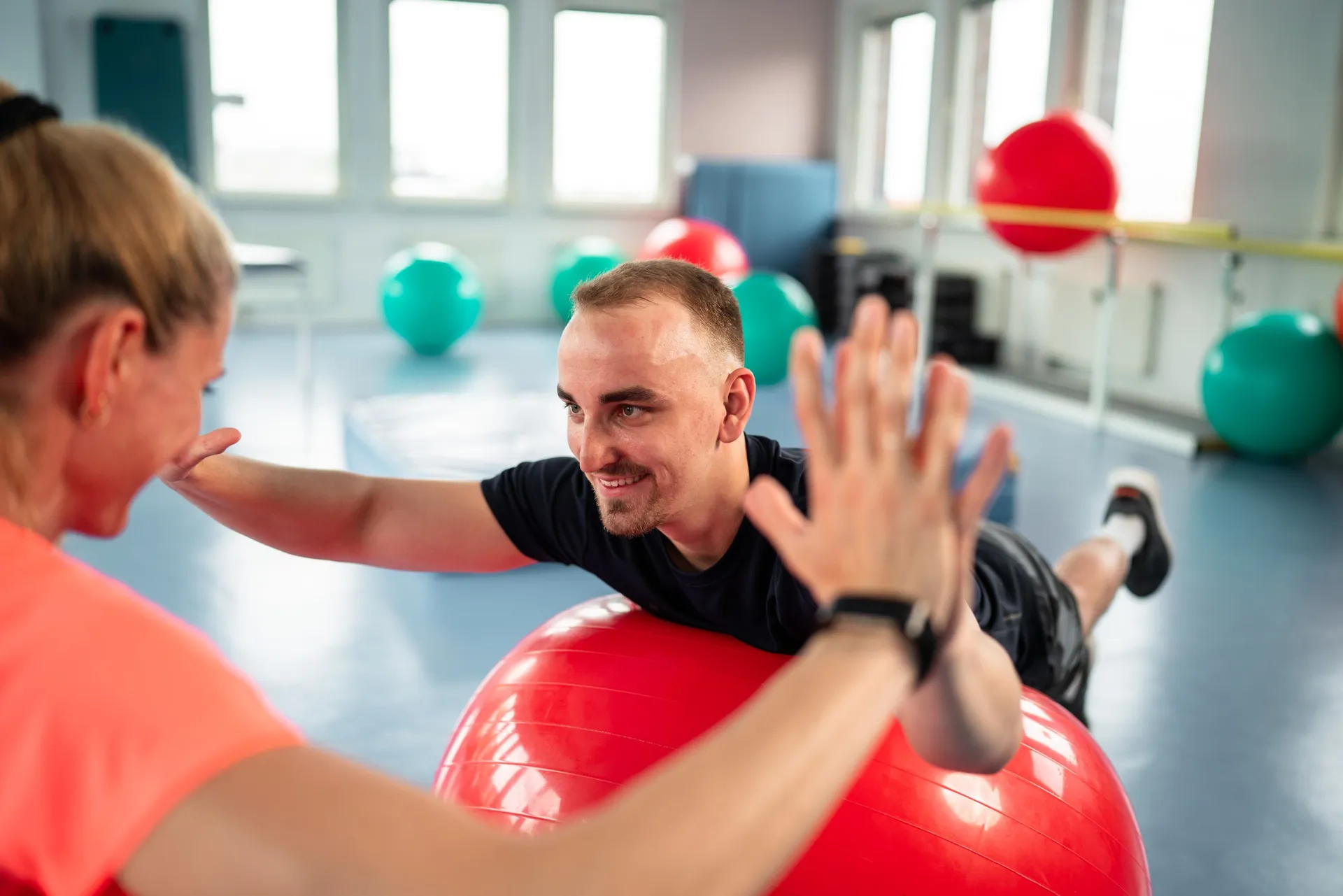 Mann liegt auf einem Gymnastikball und übt mit einer Trainerin im Fitnessraum.