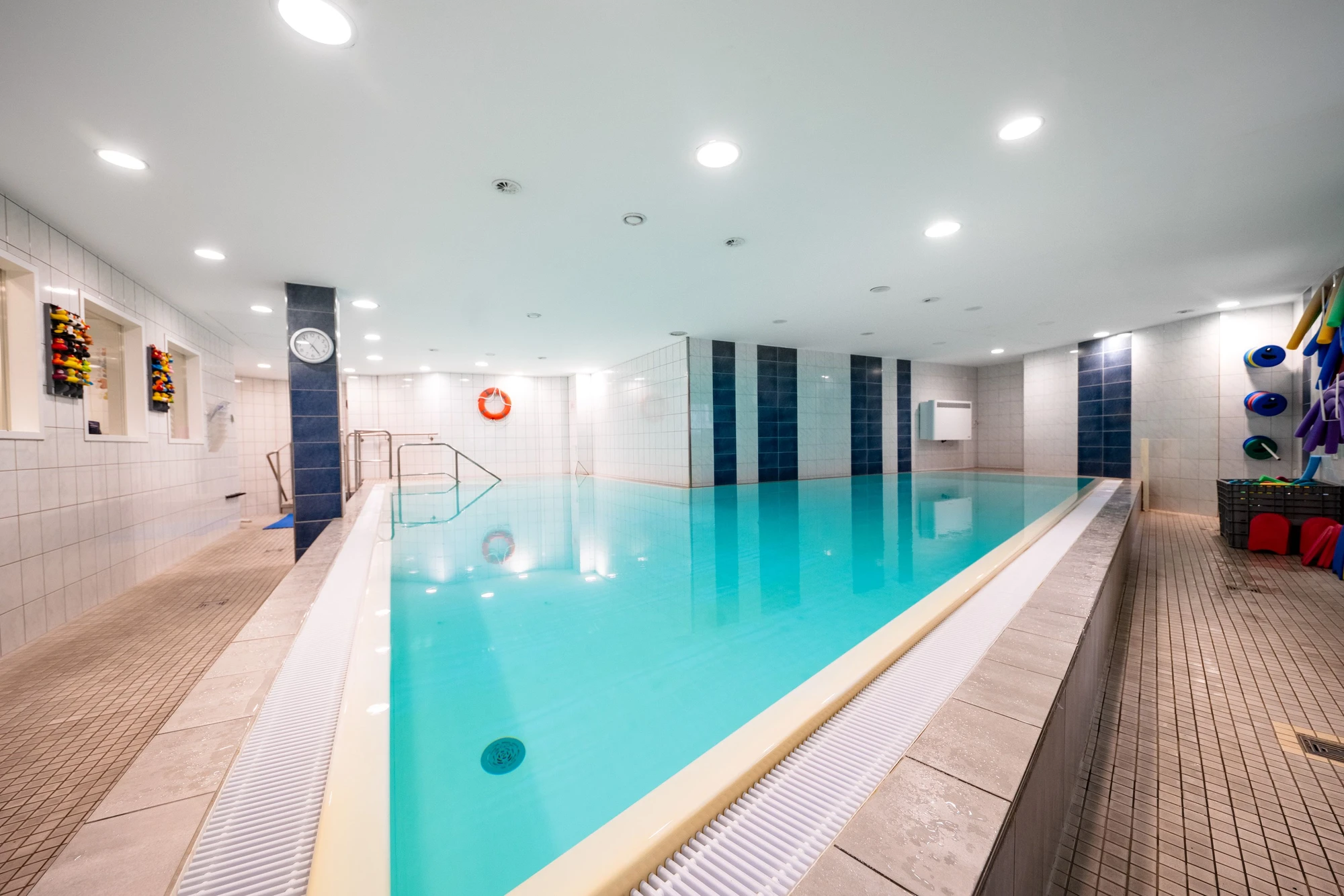 Heller Innenpool mit blauer Wasseroberfläche, Treppe und bunten Schwimmhilfen an der Wand.