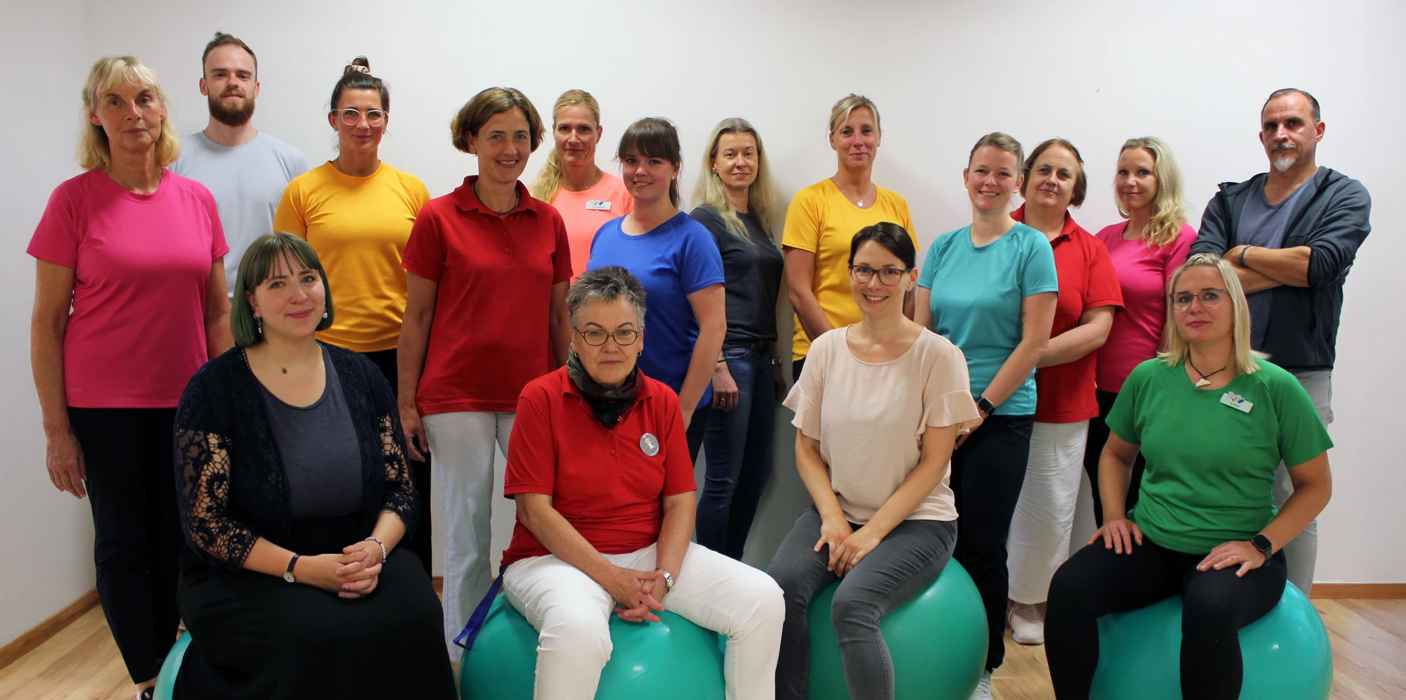 Gruppenfoto eines Teams in bunten T-Shirts, sitzend und stehend auf Gymnastikbällen.