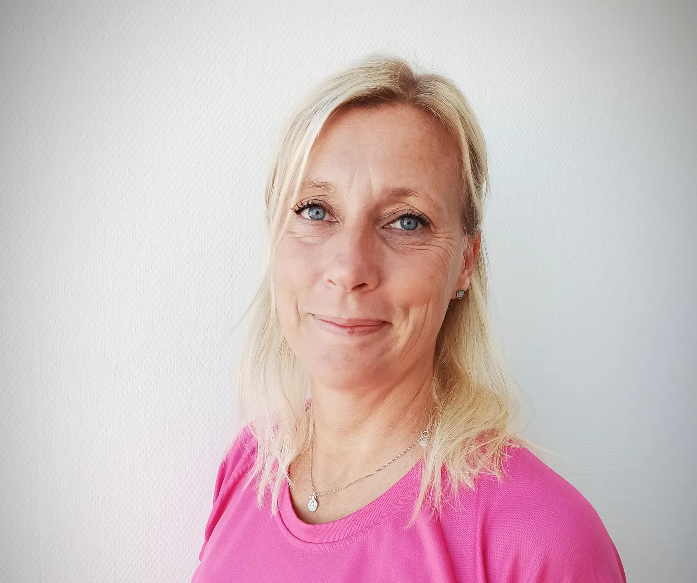 Frau mit blonden Haaren und blauen Augen lächelt in einem pinken Shirt vor weißem Hintergrund.